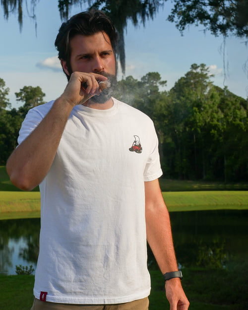 Man in a white t-shirt with a logo outdoors with cigar