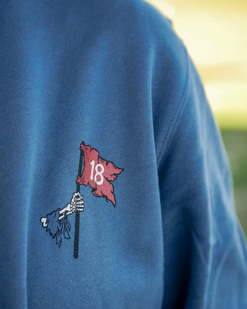 Close-up of a blue sweatshirt featuring a small embroidered graphic on the chest of a skeleton hand holding a red flag with the number 18.