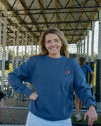 A smiling woman stands outdoors behind a chain-link fence wearing a navy blue crewneck sweatshirt with a small red and white embroidered logo on the left chest, paired with white pants.