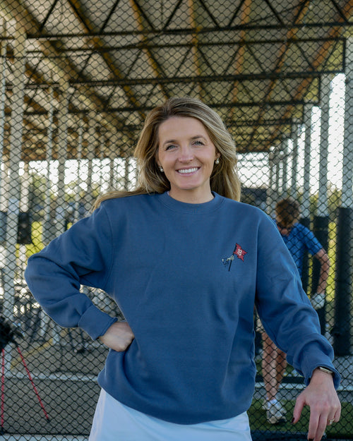 A smiling woman stands outdoors behind a chain-link fence wearing a navy blue crewneck sweatshirt with a small red and white embroidered logo on the left chest, paired with white pants.