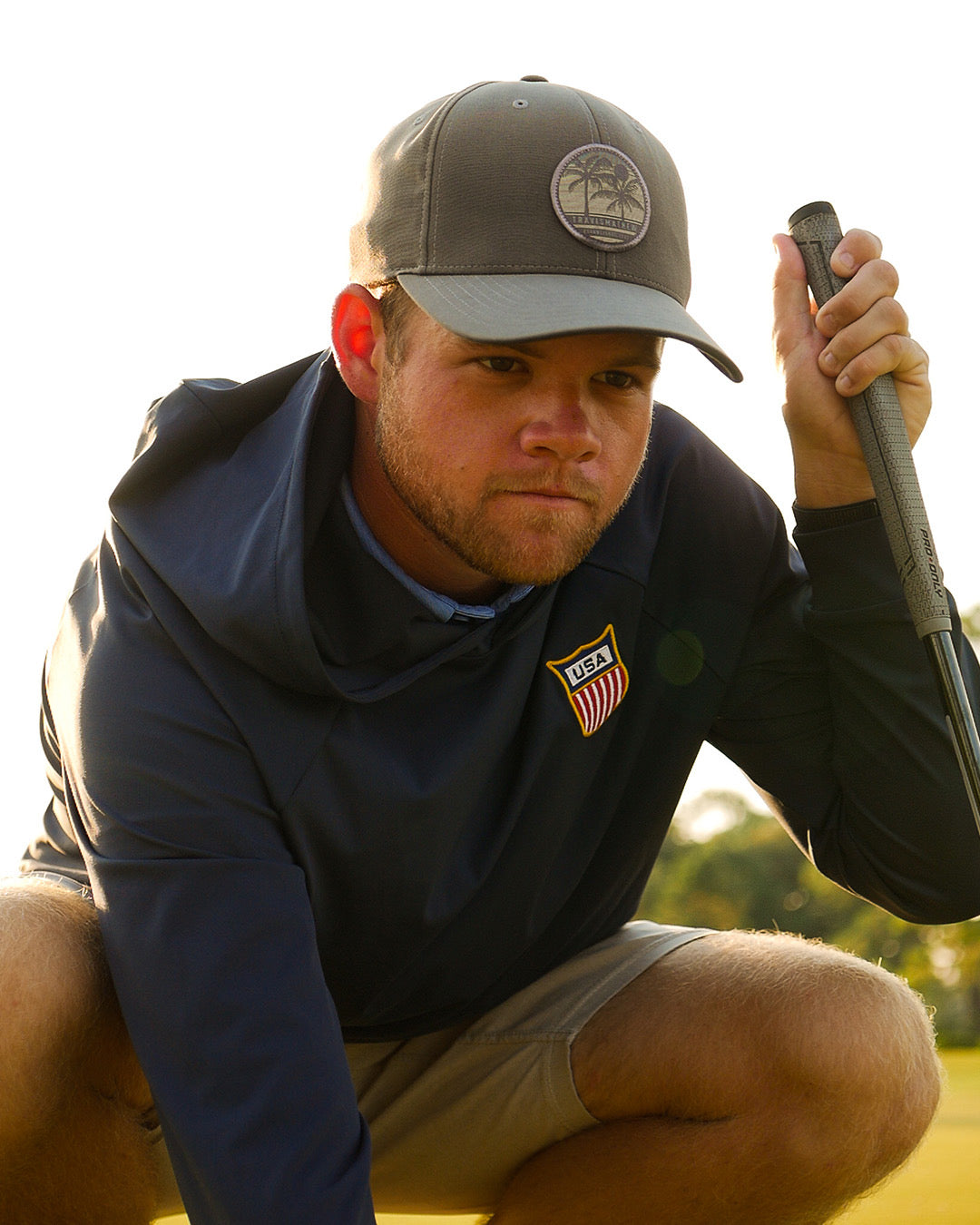 Man in a navy feather and hide golf hoodie with a USA flag patch holding a golf club