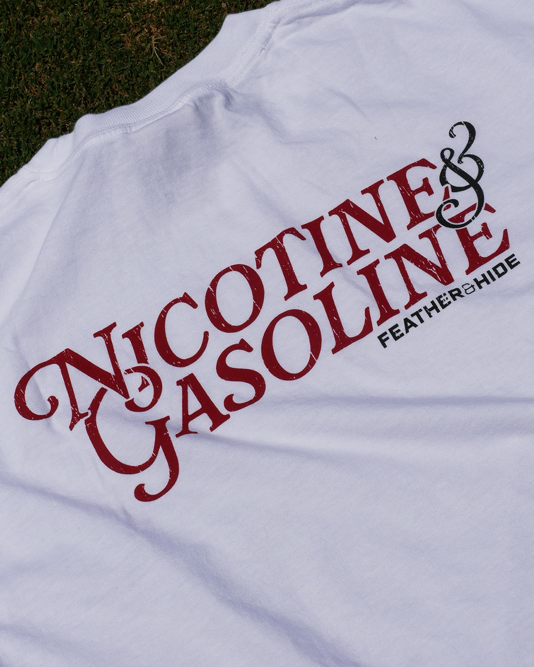 White t-shirt with red and black text 'Nicotine & Gasoline' on a grassy background
