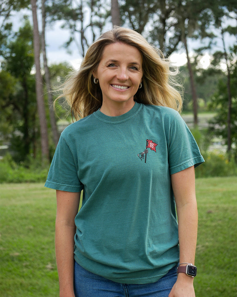 A smiling blonde woman wearing a teal green t-shirt with a small red and black flag graphic on the left chest, standing outdoors in a grassy park with trees in the background.