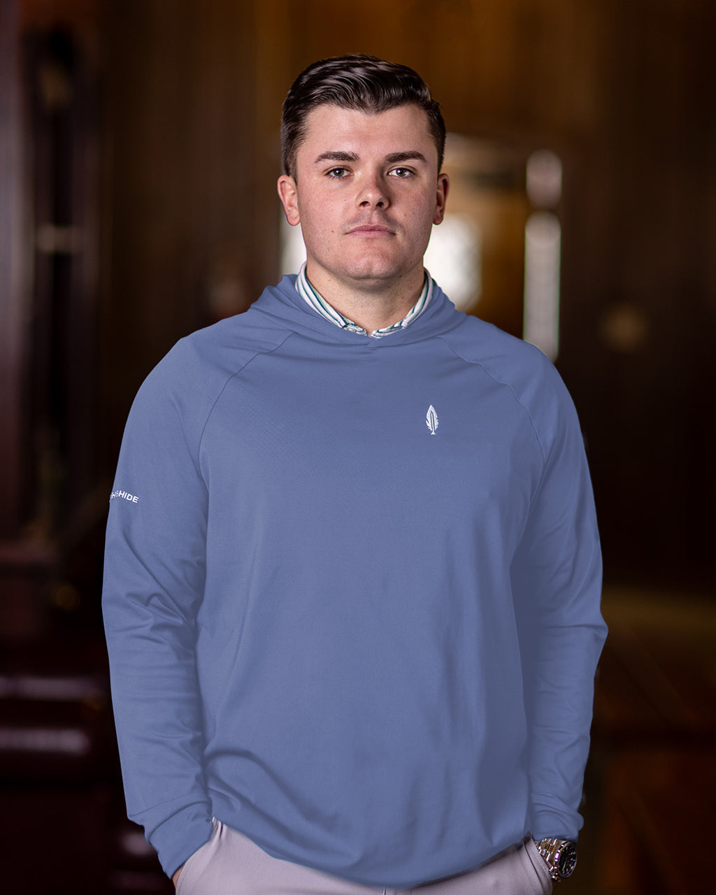 A man wearing a pale blue hoodie with the feather and hide logo on the chest, standing indoors.