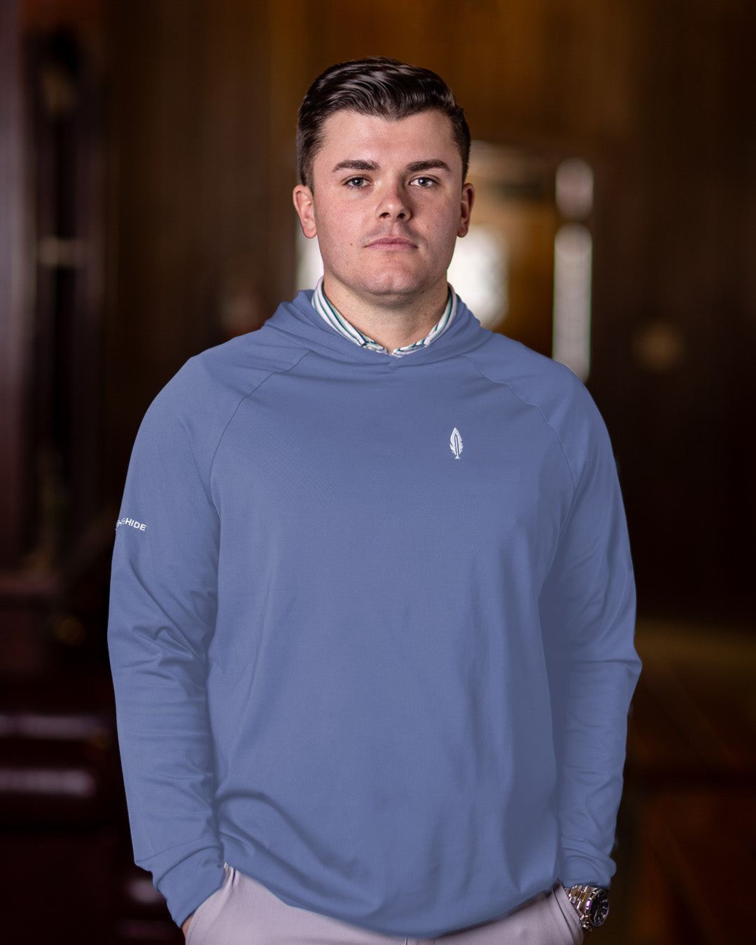 A man wearing a pale blue hoodie with the feather and hide logo on the chest, standing indoors.