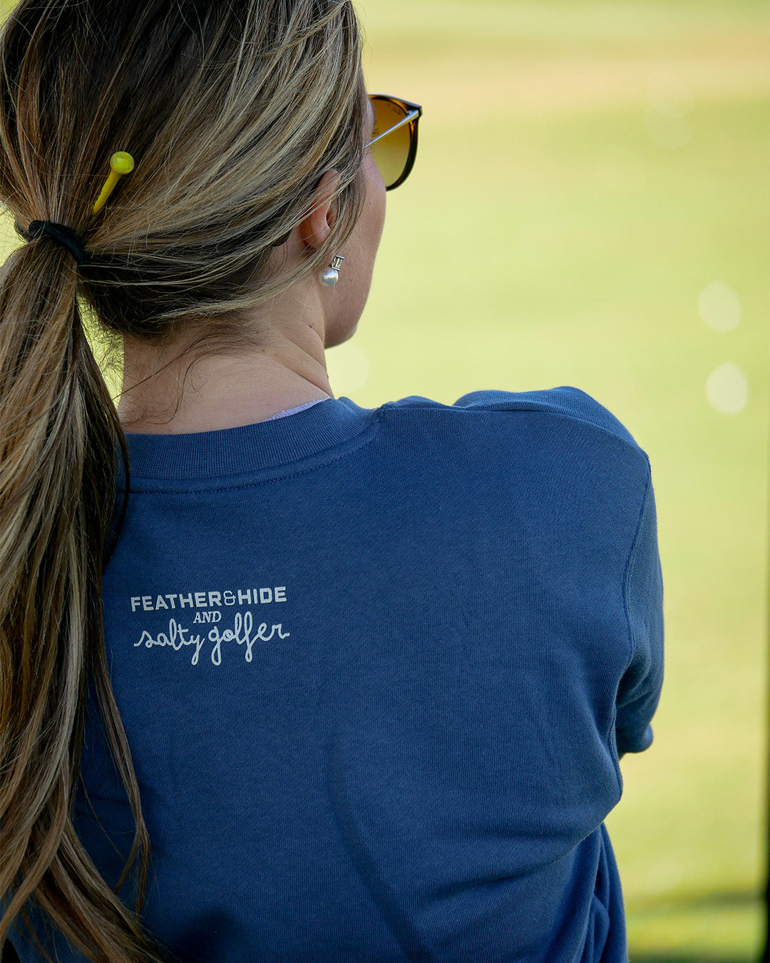 A woman viewed from behind wearing a navy blue t-shirt with 'FEATHERSHIDE and salty gaffer' printed in white on the back, set against a blurred green outdoor background.