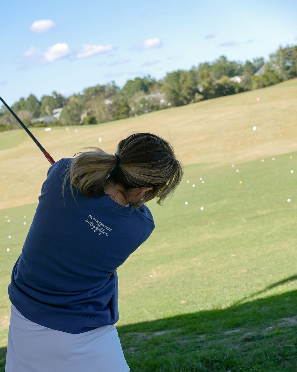 A woman in a navy blue polo shirt and white skirt swings a golf club on a sunny day at a golf course, with a grassy fairway and trees in the background.